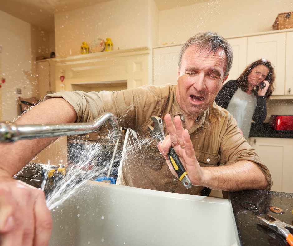 A man getting sprayed by his kitchen sink in a plumbing emergency as his wife calls a plumber in the background